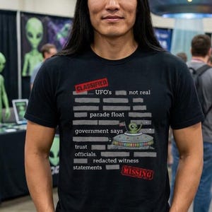 May include: Black t-shirt with the text "CLASSIFIED... UFO's not real" and a graphic of a UFO with an alien. The shirt also has the words "parade float", "government says", "trust officials", "redacted witness statements", and "MISSING".