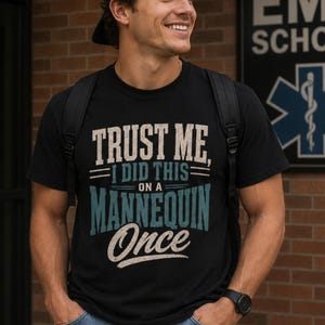 May include: Black t-shirt with the text "TRUST ME, I DID THIS ON A MANNEQUIN Once" in white and teal lettering. The shirt is worn by a person with a black backpack and watch.