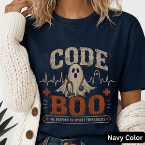 May include: A navy blue t-shirt featuring a graphic of a ghost wearing a stethoscope. The text reads CODE BOO and WE RESPOND TO SPOOKY EMERGENCIES. The design includes a heartbeat line, two orange plus signs, and a distressed, vintage-style texture.
