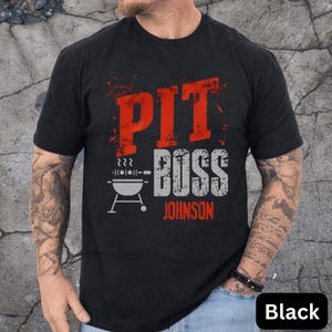 May include: Black t-shirt with the words "PIT BOSS" in red and gray, and the name "JOHNSON" in red. A graphic of a grill is below the words. The shirt is short-sleeved.