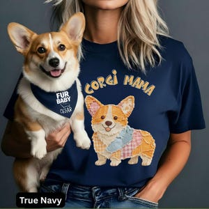 May include: A navy blue t-shirt with the words "CORGI MAMA" above an embroidered corgi design. A corgi dog wearing a navy blue bandana that says "FUR BABY CESAR" is held in front of the shirt.