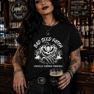 May include: Black t-shirt with a white graphic of a spooky jack-o'-lantern and the words "BAD SEED PATCH" and "LOCALLY GROWN TROUBLE". The shirt is worn by a person holding a glass of dark beer.