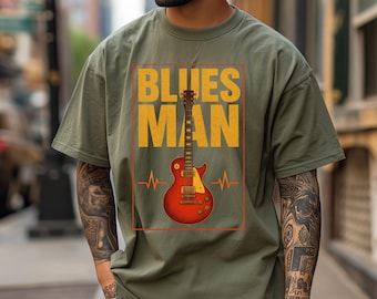 May include: Moss green t-shirt with a graphic that reads "BLUES MAN" in yellow block letters. A red electric guitar is centered below the text, with a heartbeat-like design on either side. The shirt is a relaxed fit.