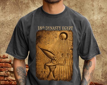May include: Graphite gray t-shirt featuring a detailed graphic of an ancient Egyptian figure with the text "18th DYNASTY EGYPT". The graphic is in shades of brown and gold, with hieroglyphs and sun rays.