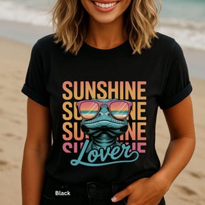 May include: Black t-shirt with a graphic of a lizard wearing sunglasses, the text "SUNSHINE LOVER" in a colorful retro font. The shirt is a classic crew neck style.