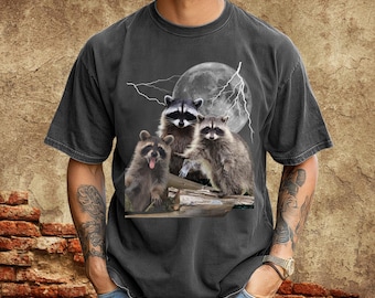 May include: Graphite-colored t-shirt featuring a graphic of three raccoons with a full moon and lightning in the background. The raccoons are depicted in a realistic style, with detailed fur and facial expressions. The shirt is a casual, short-sleeved style.
