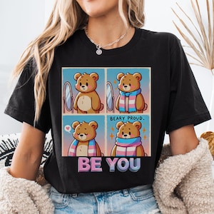 May include: Black t-shirt with a cartoon illustration of a bear wearing a transgender pride scarf. The text "Beary Proud" and "Be You" are printed on the shirt.