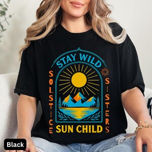 May include: Black t-shirt with a graphic design featuring a yellow sun, mountains, and the text "Stay Wild Solstice Sisters Sun Child" in blue and orange. The shirt has a crew neck and short sleeves.