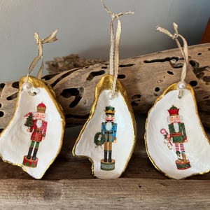 May include: Three handmade Christmas ornaments shaped like oyster shells with gold trim. Each ornament features a different nutcracker design: one in red, one in blue, and one in green.