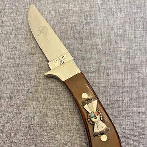 May include: A brown handled knife with a silver blade. The blade has the words "Arapaho" and "Case XX USA" etched into it. The handle has a decorative inlay of turquoise, mother of pearl, and silver.