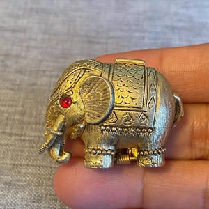 May include: A gold-tone elephant brooch with red rhinestone eyes. The elephant is detailed with intricate carvings and bead accents.