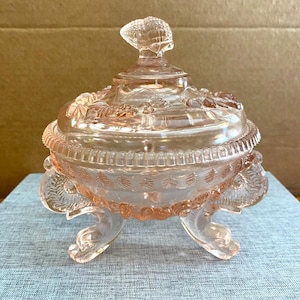 May include: A pink, antique glass candy dish with a lid. The dish is round with a decorative rim and sits on three curved feet. The lid has a seashell-shaped handle. The glass has a delicate, translucent appearance.