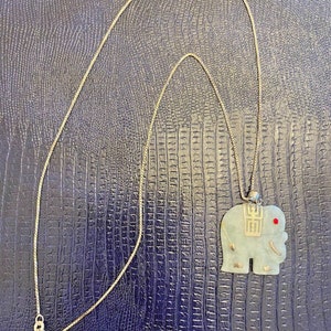 May include: A jade green elephant pendant with gold accents and a red stone for an eye hangs on a gold chain necklace.
