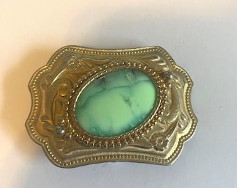 May include: A gold-toned belt buckle with an oval-shaped green stone in the center. The buckle has a decorative design around the stone and on the edges.