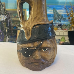 May include: A wooden carved pirate head bottle with a black eye patch and a black hat. The bottle is brown and has a textured surface.