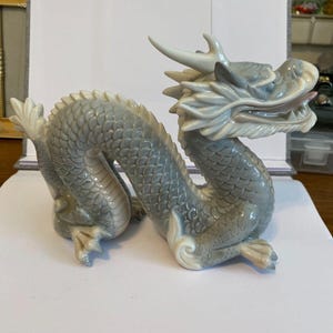 May include: A gray porcelain dragon figurine with a white underside. The dragon is curled up with its head raised and its mouth open. The figurine is signed "Geshimin" on the bottom.