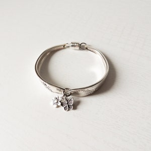 May include: A silver bracelet made from a spoon, featuring a floral and butterfly charm. The bracelet has a magnetic clasp and is displayed on a white surface. The bracelet is approximately 7 inches in circumference.