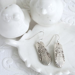 May include: A pair of silver earrings with a floral design. The earrings are hanging on a white ceramic dish.