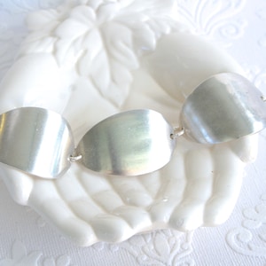 May include: A silver bracelet with three large, smooth, oval-shaped links. The links are connected by small silver rings. The bracelet is displayed on a white, hand-shaped ceramic holder, against a white background.