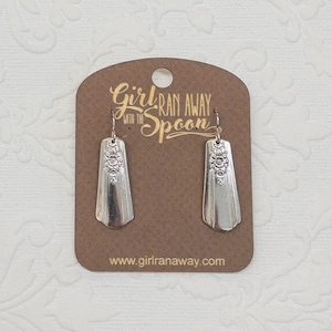 May include: Silver-colored earrings with a floral design, displayed on a brown card. The earrings are rectangular with a decorative floral pattern at the top. The card has the text "Girl Ran Away with the Spoon" and the website address.