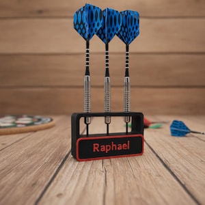Könnte beinhalten: Drei Dartpfeile mit blau-schwarzen Flights werden in einem schwarz-roten Halter präsentiert. Der Halter trägt den Namen "Raphael". Die Darts liegen auf einer Holzoberfläche.