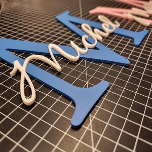 May include: Blue and white custom name cutouts. The name "Michel" is displayed in a cursive font, with the name in white and the base in blue. The cutouts are on a black grid surface.