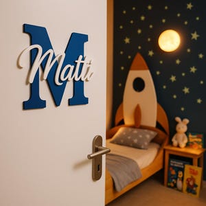 May include: A blue wooden letter "M" with the name "Matti" in white script, mounted on a white door. The room features a rocket-shaped bed, a nightstand, and a wall decorated with stars and a moon-shaped light.