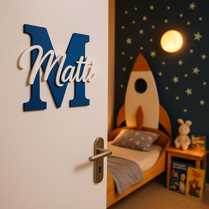 May include: A blue wooden letter "M" with the name "Matti" in white script, mounted on a white door. The room features a rocket-shaped bed, a nightstand, and a wall decorated with stars and a moon-shaped light.