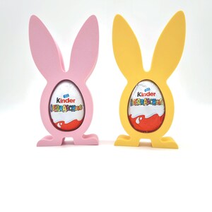 Könnte beinhalten: Zwei Osterhasen-förmige Halter, einer rosa und einer gelb, die jeweils ein Kinder Überraschung-Schokoladenei halten. Die Eier sind in Folie verpackt, auf der "Kinder Überraschung" steht.