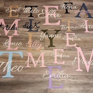 May include: Assorted wooden letters and names in various colors, including pink, blue, black, and gold. Names like "Emil", "Mila", and "Theo" are displayed in cursive script. The letters are arranged on a wooden surface.