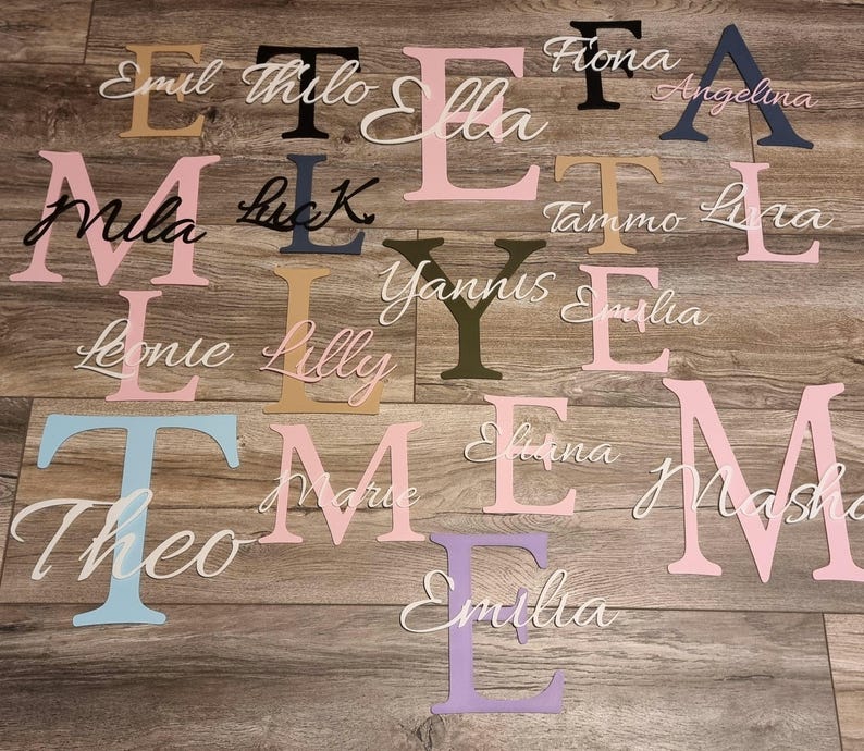 May include: Assorted wooden letters and names in various colors, including pink, blue, black, and gold. Names like "Emil", "Mila", and "Theo" are displayed in cursive script. The letters are arranged on a wooden surface.