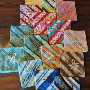 May include: A collection of square, crocheted dishcloths arranged in a cluster. Each cloth features diagonal stripes in various color combinations, including blue, green, yellow, pink, brown, and white, with a small matching string tied in a bow.