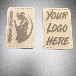 Puede incluir: Dos parches rectangulares con bordes cosidos. Un parche presenta una figura de samurái y el texto "RHONIN CREATES". El otro parche tiene el texto "YOUR LOGO HERE". Ambos parches son de color beige con diseños marrón oscuro.