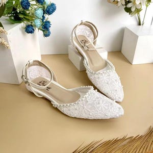 Könnte beinhalten: Ein Paar weiße, flache Schuhe mit Knöchelriemen. Die Schuhe sind mit einem zarten floralen Spitzenmuster bedeckt und mit kleinen Perlen an den Rändern verziert. Der Hintergrund ist ein neutrales Beige.