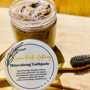 May include: A glass jar of brown mineralizing toothpaste with a white label that reads "Cosmic North Apothecary Mineralizing Toothpaste" and lists the ingredients: bentonite clay, dolomite, sodium carbonate, xylitol, MCT oil, calcium hydroxapatite, silica, trace minerals, potassium, celtic salt, nascent iodine, clove extract, wintermint eo.