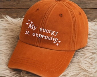 May include: An orange baseball cap with the phrase "My energy is expensive" in white script. The hat is made of a soft material and has a curved brim. The hat is resting on a white fluffy surface.