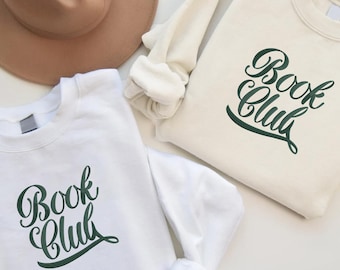 May include: Two white sweatshirts with the text "Book Club" in green cursive lettering. The sweatshirts are folded and laid out on a white surface.