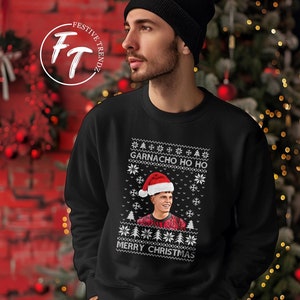 Könnte beinhalten: Schwarzes Sweatshirt mit festlichem Design. Das Design enthält den Text "GARNACHO HO HO" und "MERRY CHRISTMAS", ein Porträt einer Person mit Weihnachtsmütze, Schneeflocken und Weihnachtsbäumen. Das Sweatshirt wird von einer Person mit schwarzer Mütze getragen.