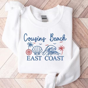 May include: White sweatshirt with the text "Cousins Beach EAST COAST" in blue script. The design includes a seashell, waves, palm trees, and a red and white floral element. A casual, beach-themed apparel item.