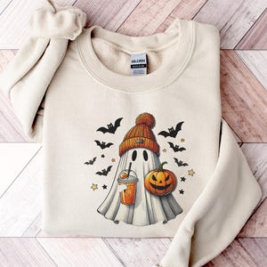 May include: A cream-coloured sweatshirt with a Halloween graphic. The design shows a ghost in an orange beanie, holding a drink, with bats, a jack-o'-lantern, and stars. The sweatshirt is folded on a wooden surface.