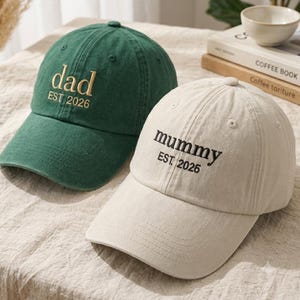 Puede incluir: Una gorra de béisbol verde oscuro con el texto bordado dad EST. 2026 en dorado y una gorra beige con el texto bordado mummy EST. 2026 en negro. Ambas gorras cuentan con una corona estructurada, una visera curva y ojales cosidos.