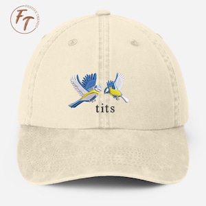 Peut inclure: Une casquette de baseball beige avec une image brodée de deux mésanges bleues se faisant face. Le texte "tits" est brodé sous les oiseaux.