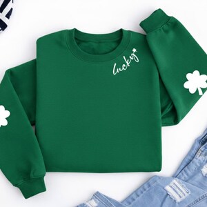 Puede incluir: Una sudadera verde con gráficos de tréboles blancos en las mangas y la palabra "Lucky" en escritura cursiva. La sudadera se combina con unos vaqueros azules y un sombrero a rayas.