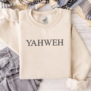 May include: A beige crewneck sweatshirt featuring the word YAHWEH embroidered in dark gray thread across the center of the chest. The garment has long sleeves with ribbed cuffs and a ribbed neckline, presented with a pair of gray denim jeans.