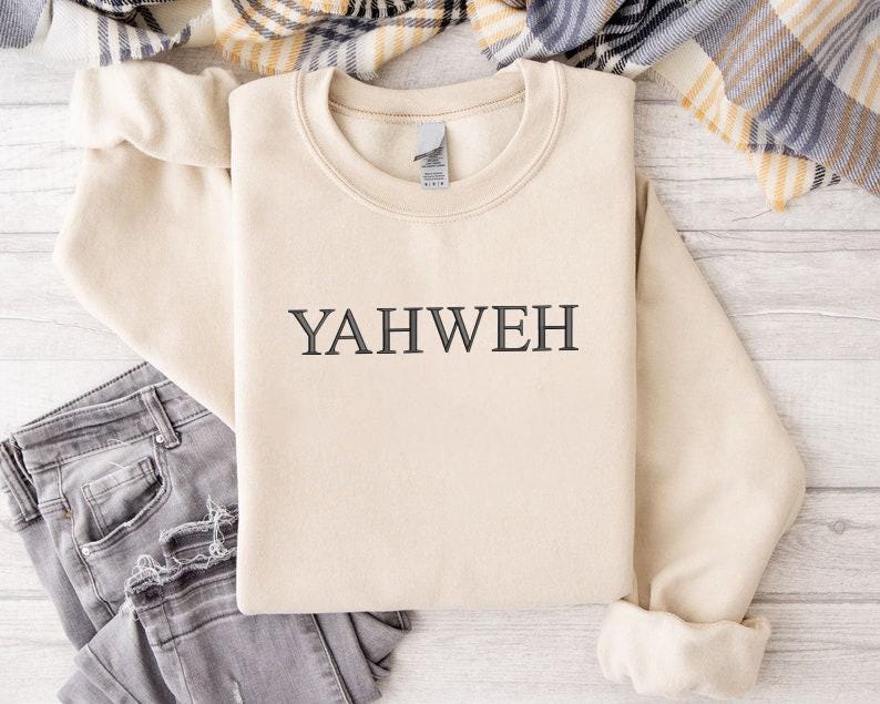 May include: A beige crewneck sweatshirt featuring the word YAHWEH embroidered in dark gray thread across the center of the chest. The garment has long sleeves with ribbed cuffs and a ribbed neckline, presented with a pair of gray denim jeans.