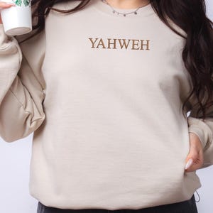 May include: A beige crewneck sweatshirt featuring the word YAHWEH embroidered in brown thread across the center of the chest. The garment has long sleeves with ribbed cuffs and a ribbed neckline, providing a simple and clean aesthetic design.