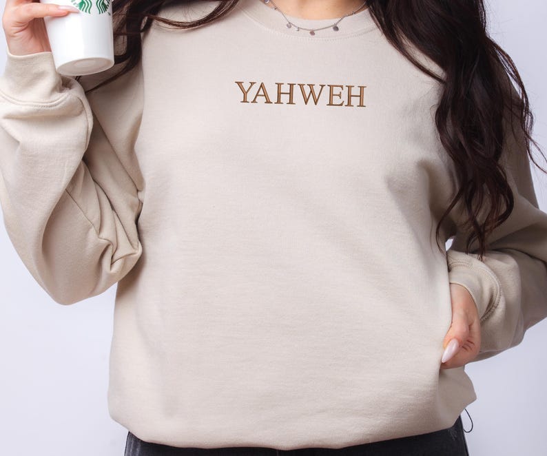 May include: A beige crewneck sweatshirt featuring the word YAHWEH embroidered in brown thread across the center of the chest. The garment has long sleeves with ribbed cuffs and a ribbed neckline, providing a simple and clean aesthetic design.