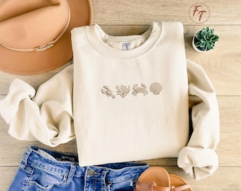 May include: Cream-colored sweatshirt with embroidered sea-themed designs, including a lobster, coral, crab, and seashell. The sweatshirt is paired with blue denim shorts, a tan hat, and sandals, creating a casual, beachy aesthetic.