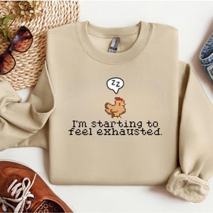 Peut inclure: Un sweat-shirt beige avec un motif de poulet pixélisé et le texte "I'm starting to feel exhausted." Le poulet a une bulle de dialogue avec "zz". Le sweat-shirt est plié sur une surface blanche.