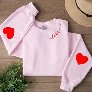 Può includere: Una felpa rosa chiaro con motivi a cuore rossi sulle maniche e la parola "love" in corsivo rosso. Questa comoda felpa è ideale per l'uso quotidiano o come regalo di San Valentino. Realizzata in tessuto morbido.